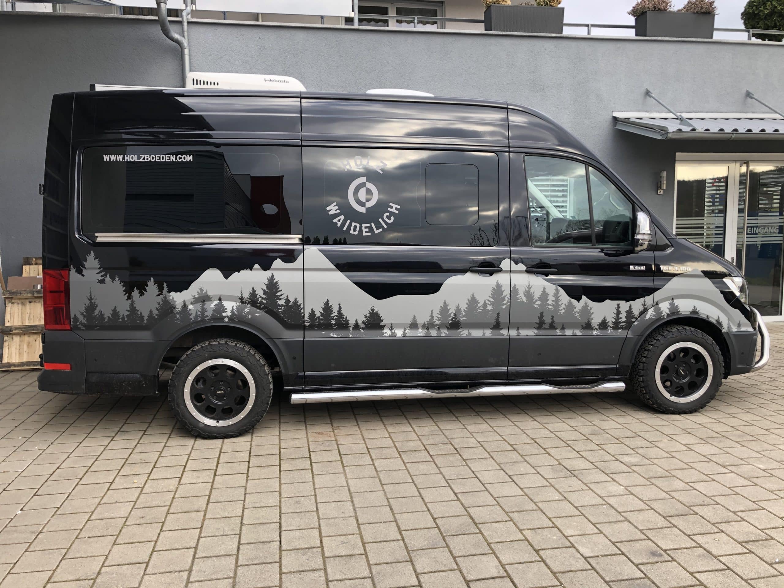 Schwarzer Transporter mit Bergsilhouette und Holz-Werbung